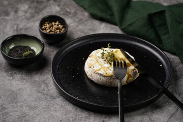 Oven baked camembert cheese with black pepper, sesame seeds on black plate, grey concrete surface. Homemade grilled brie with thyme, dark table setting