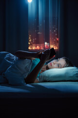 Naklejka premium Young woman using smartphone in bed in her cozy bedroom at night.