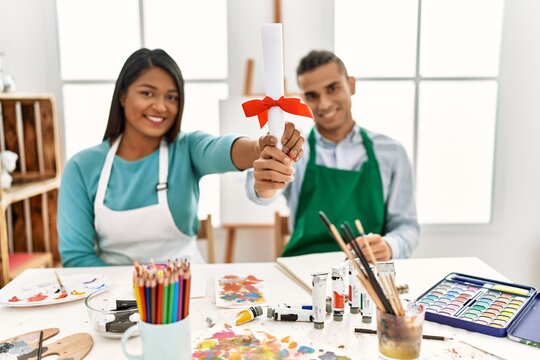 Young latin painter couple smiling happy holding diploma sitting on the table at art studio