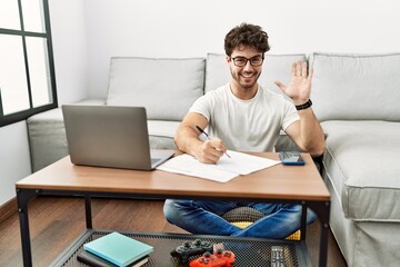 Hispanic man doing papers at home waiving saying hello happy and smiling, friendly welcome gesture