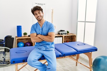 Young hispanic man wearing physio therapist uniform with arms crossed gesture at clinic