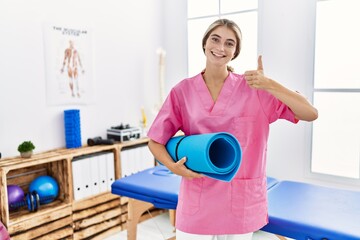 Young physiotherapist woman working at pain recovery clinic holding yoga mat smiling happy and positive, thumb up doing excellent and approval sign