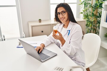 Young hispanic woman wearing doctor uniform holding pills working at clinic