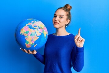 Young blonde woman holding world ball smiling with an idea or question pointing finger with happy...