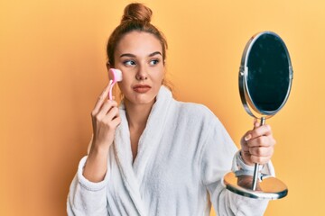 Young blonde woman wearing robe holding makeup remover brush relaxed with serious expression on face. simple and natural looking at the camera.