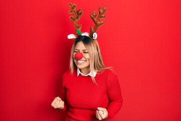 Beautiful hispanic woman wearing deer christmas hat and red nose very happy and excited doing winner gesture with arms raised, smiling and screaming for success. celebration concept.