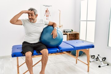 Fototapeta na wymiar Middle age hispanic man at pain recovery clinic holding pilates ball very happy and smiling looking far away with hand over head. searching concept.
