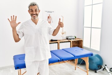 Fototapeta na wymiar Middle age hispanic therapist man working at pain recovery clinic showing and pointing up with fingers number six while smiling confident and happy.