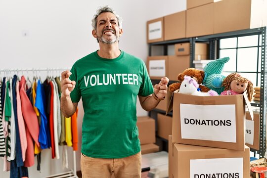 Middle age hispanic man wearing volunteer t shirt at donations stand gesturing finger crossed smiling with hope and eyes closed. luck and superstitious concept.