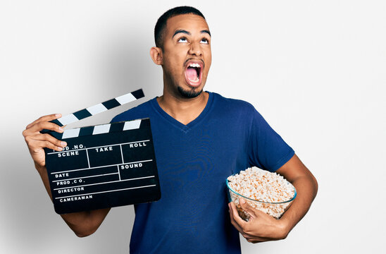 Young African American Man Eating Popcorn Holding Film Clapboard Angry And Mad Screaming Frustrated And Furious, Shouting With Anger Looking Up.