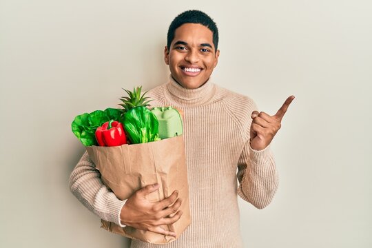 Young handsome hispanic man holding paper bag with groceries smiling happy pointing with hand and finger to the side