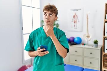 Young caucasian physio man using smartphone at medical clinic serious face thinking about question with hand on chin, thoughtful about confusing idea