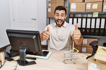 Handsome hispanic man working at small business commerce approving doing positive gesture with hand, thumbs up smiling and happy for success. winner gesture.