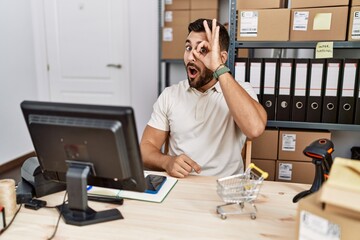 Handsome hispanic man working at small business commerce doing ok gesture shocked with surprised face, eye looking through fingers. unbelieving expression.