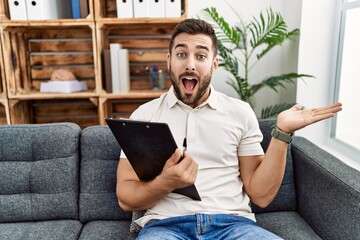 Handsome hispanic man holding clipboard working at psychology clinic pointing aside with hands open...
