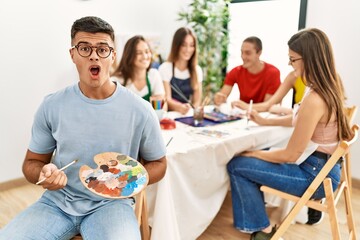 Young artist man drawing at art studio scared and amazed with open mouth for surprise, disbelief face