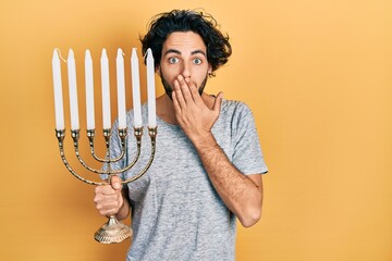 Handsome hispanic man holding menorah hanukkah jewish candle covering mouth with hand, shocked and...