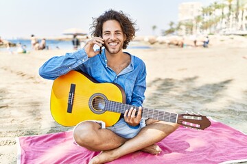 Young hispanic man playing guitar and talking on the smartphone sitting on sand at the beach.