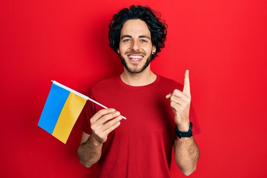 Handsome hispanic man wearing ukraine flag smiling with an idea or question pointing finger with happy face, number one