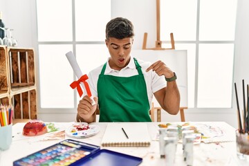 Young hispanic man at art studio holding degree pointing down with fingers showing advertisement,...