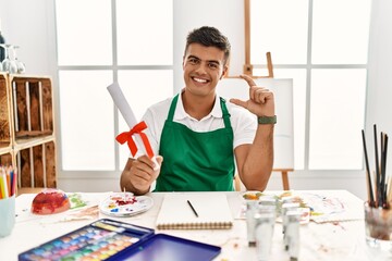 Young hispanic man at art studio holding degree smiling and confident gesturing with hand doing small size sign with fingers looking and the camera. measure concept.