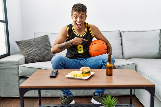 Young handsome hispanic man holding basketball ball cheering tv game smiling happy pointing with hand and finger