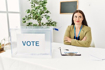 Young brunette woman sitting at election table with voting ballot smiling looking to the side and...