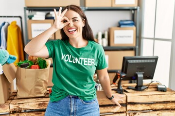 Young brunette woman wearing volunteer t shirt at donations stand doing ok gesture with hand smiling, eye looking through fingers with happy face.