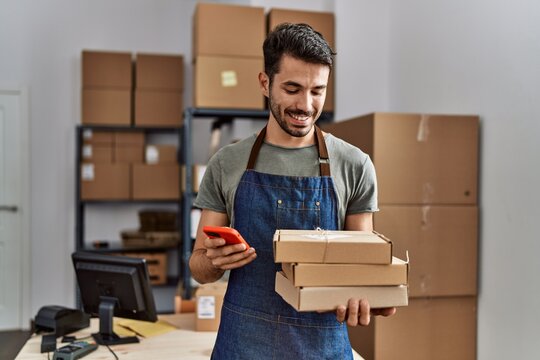 Young hispanic man business worker using smartphone holding packages at storehouse