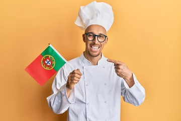 Fotobehang Bald man with beard wearing professional cook apron holding portugal flag smiling happy pointing with hand and finger © Krakenimages.com