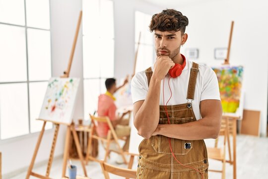 Young hispanic man at art studio looking confident at the camera with smile with crossed arms and hand raised on chin. thinking positive.