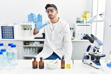 Young hispanic man working at scientist laboratory with open hand doing stop sign with serious and confident expression, defense gesture