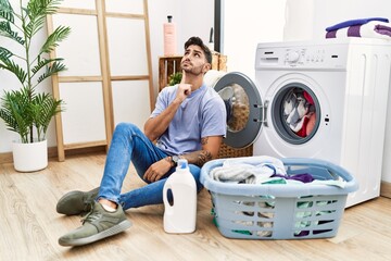 Young hispanic man putting dirty laundry into washing machine thinking concentrated about doubt with finger on chin and looking up wondering