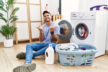 Young hispanic man putting dirty laundry into washing machine showing middle finger doing fuck you...