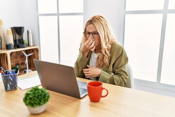 Beautiful blonde woman working at the office with laptop smelling something stinky and disgusting, intolerable smell, holding breath with fingers on nose. bad smell - obrazy, fototapety, plakaty