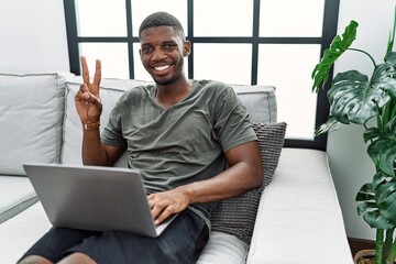 Young african american man using laptop at home sitting on the sofa smiling with happy face winking...