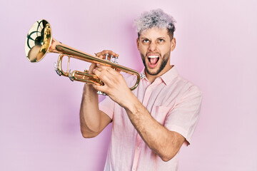 Young hispanic man with modern dyed hair playing trumpet angry and mad screaming frustrated and...