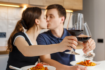 Romantic dinner at home. Happy couple kissing, clinking glasses and eating bolognese pasta with wine at kitchen. Concept of domestic lifestyle, happy marriage and togetherness. Focus on foreground.