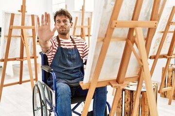 Young handsome man with beard at art studio sitting on wheelchair with open hand doing stop sign with serious and confident expression, defense gesture