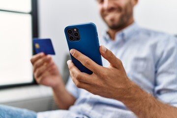 Young hispanic man using smartphone and credit card sitting on the sofa at home.