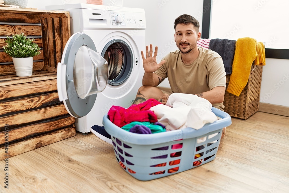 Poster Young handsome man putting dirty laundry into washing machine showing and pointing up with fingers number five while smiling confident and happy.