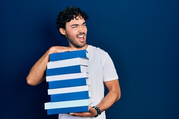 Young hispanic man holding a pile of books angry and mad screaming frustrated and furious, shouting with anger. rage and aggressive concept.