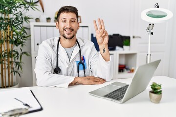 Obraz premium Young doctor working at the clinic using computer laptop showing and pointing up with fingers number three while smiling confident and happy.