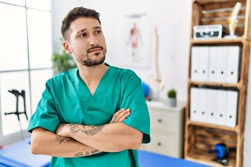 Young physiotherapist man working at pain recovery clinic looking to the side with arms crossed convinced and confident