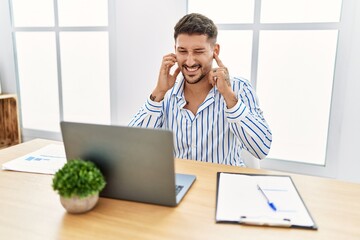 Young handsome man with beard working at the office using computer laptop covering ears with...