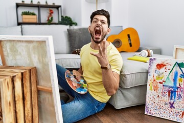 Young man with beard painting canvas at home crazy and mad shouting and yelling with aggressive...