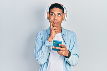 Young african american guy using smartphone wearing headphones serious face thinking about question...