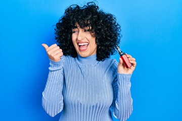 Young middle east woman holding electronic cigarette pointing thumb up to the side smiling happy...