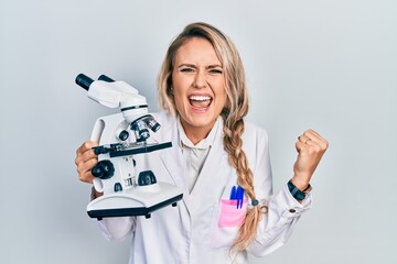 Beautiful young blonde woman holding microscope screaming proud, celebrating victory and success very excited with raised arms