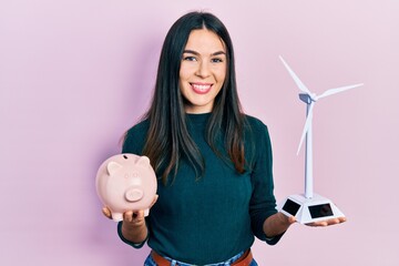 Young brunette woman holding piggy bank and wind turbine smiling with a happy and cool smile on...
